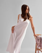 Woman in a white dress standing next to a chair with a floral pattern on a plain background